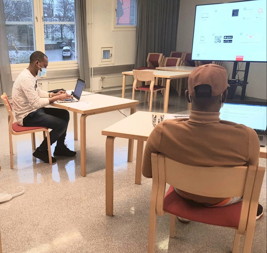 Kaksi nuorta miestä istuvat selin kuvaajaan, kumpikin oman pöydän äärellä ja maskit kasvoilla. Molemmat nuoret kirjoittavat kannettavalla tietokoneella.