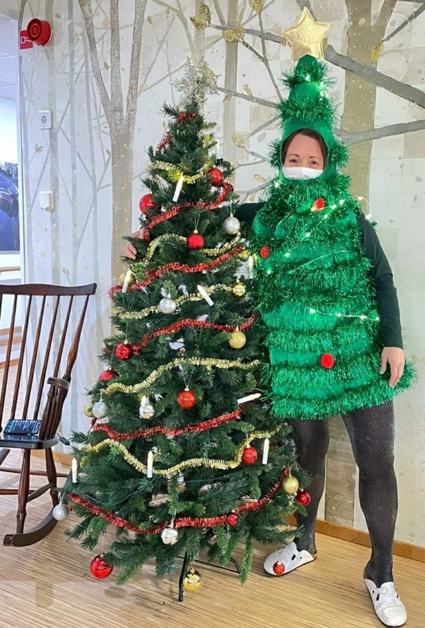 joulukuusiasuun pukeutunut henkilö poseeraa muovisen joulukuusen vieressä.