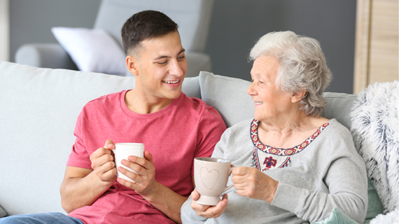 nuori mies ja vanha nainen istuvat sohvalla vierekkäin kahvikupit kädessä ja hymyilevät.