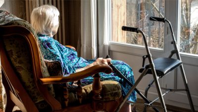 vanha nainen istuu nojatuolissa ja katsoo ulos. kädessään hänellä on kävelykeppi ja vieressä rollaattori.