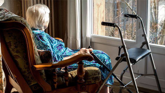 vanha nainen istuu nojatuolissa ja katsoo ulos. kädessään hänellä on kävelykeppi ja vieressä rollaattori.