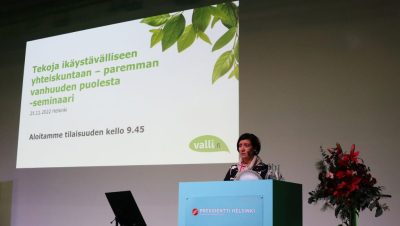 Hanna-Leena Ojalainen puhujapöntössä, taustalla heijastettuna seminaarin nimi Tekoja ikäystävälliseen yhteiskuntaan - paremman vanhuuden puolesta -seminaari