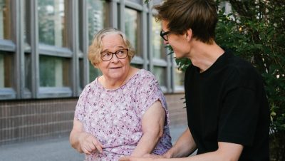 Vanhempi nainen ja nuorempi mies istuvat ulkona vierekkäin. Nainen pitää älypuhelinta kädessään.