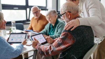 Digiraadin kuvituskuva Pöydän äärellä istuu joukko senioreita, ja heillä on älytabletit kädessään. Etualalla istuvan seniorin vieressä seisoo henkilö, joka on laskenut kätensä istuvan henkilön harteille.
