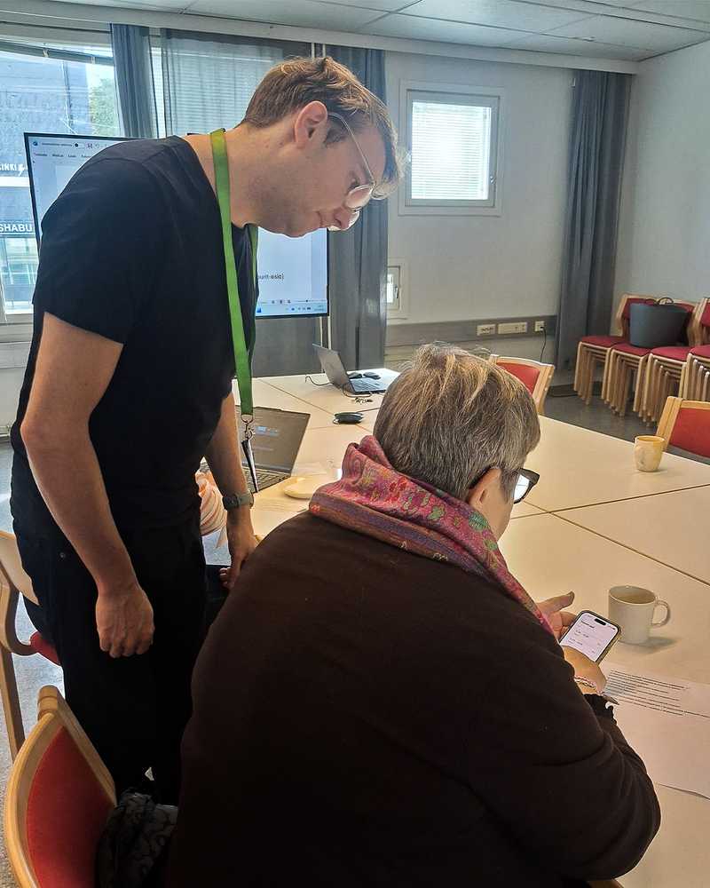 Takaapäin otettu kuva henkilöstä istumassa pöydän äärellä. Vieressä seisoo toinen henkilö, joka tarkkailee istuvan henkilön älypuhelimen käyttöä.