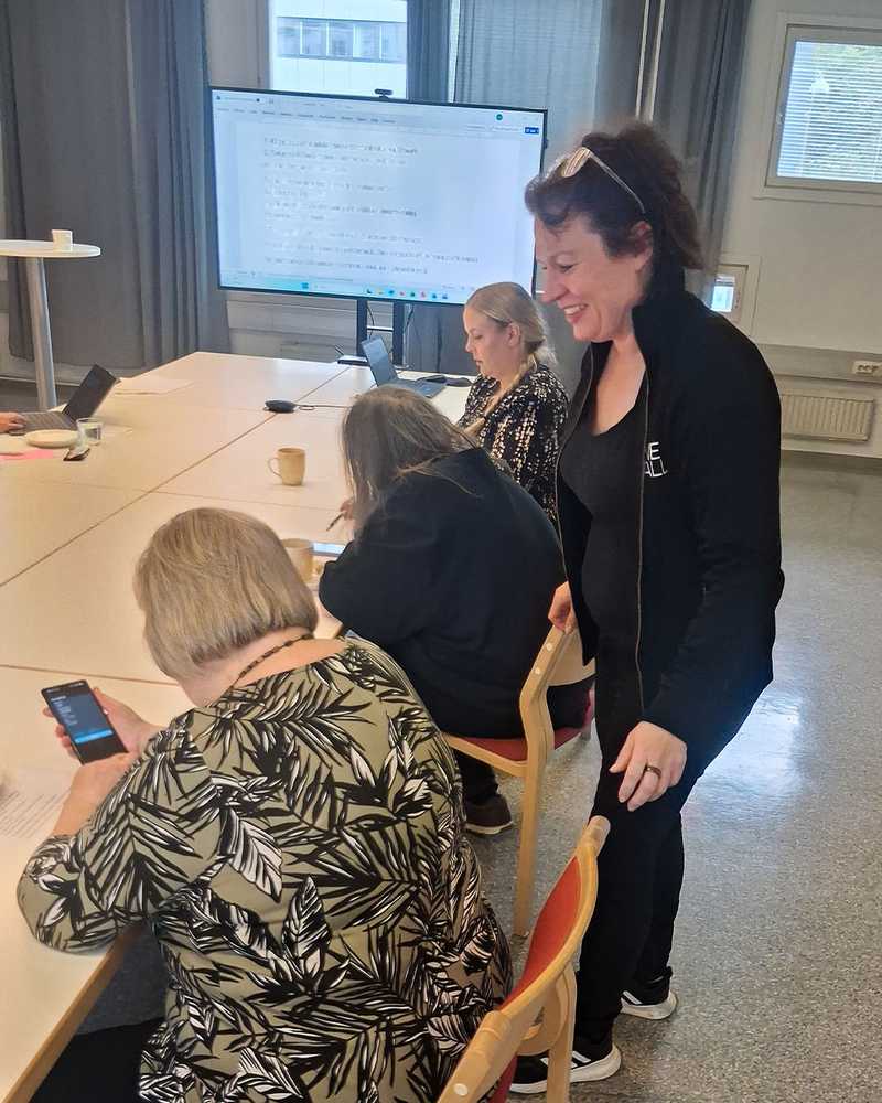 Sivusta otettu kuva kolmesta istuvasta ja yhdestä seisovasta henkilöstä. Etualalla olevan istuvan henkilön kädessä on älypuhelin.